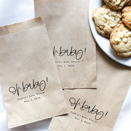 Brown paper bags with 'Oh baby!' text for a baby shower, next to a plate of cookies.