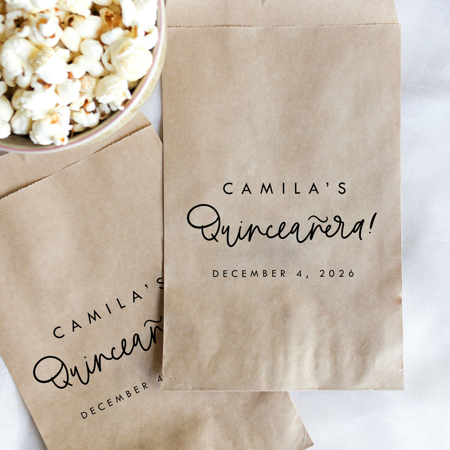 Kraft paper bags with personalized text for a Quinceanera event, next to a bowl of popcorn on a white surface.