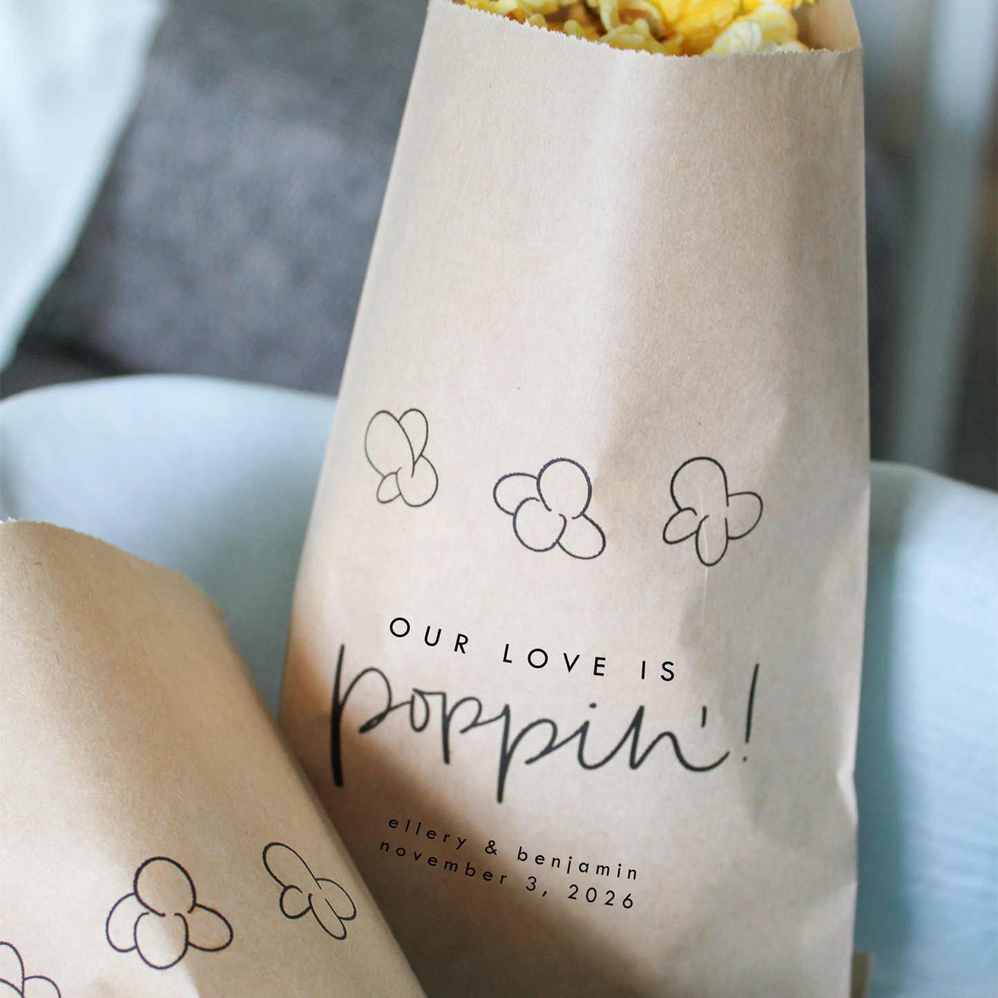 Two brown paper bags filled with popcorn, each with 'Our Love is Poppin!' text and decorative elements.