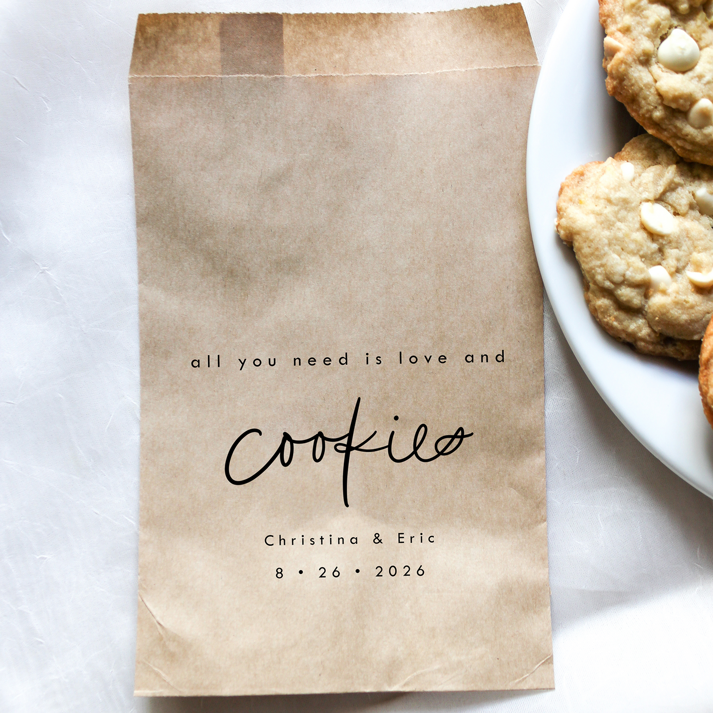 Brown paper bag with 'all you need is love and cookies' text next to a plate of cookies on a white surface