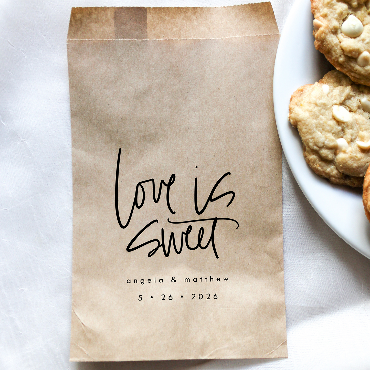 Kraft paper bag with 'Love is Sweet' text next to a plate of cookies on a white surface