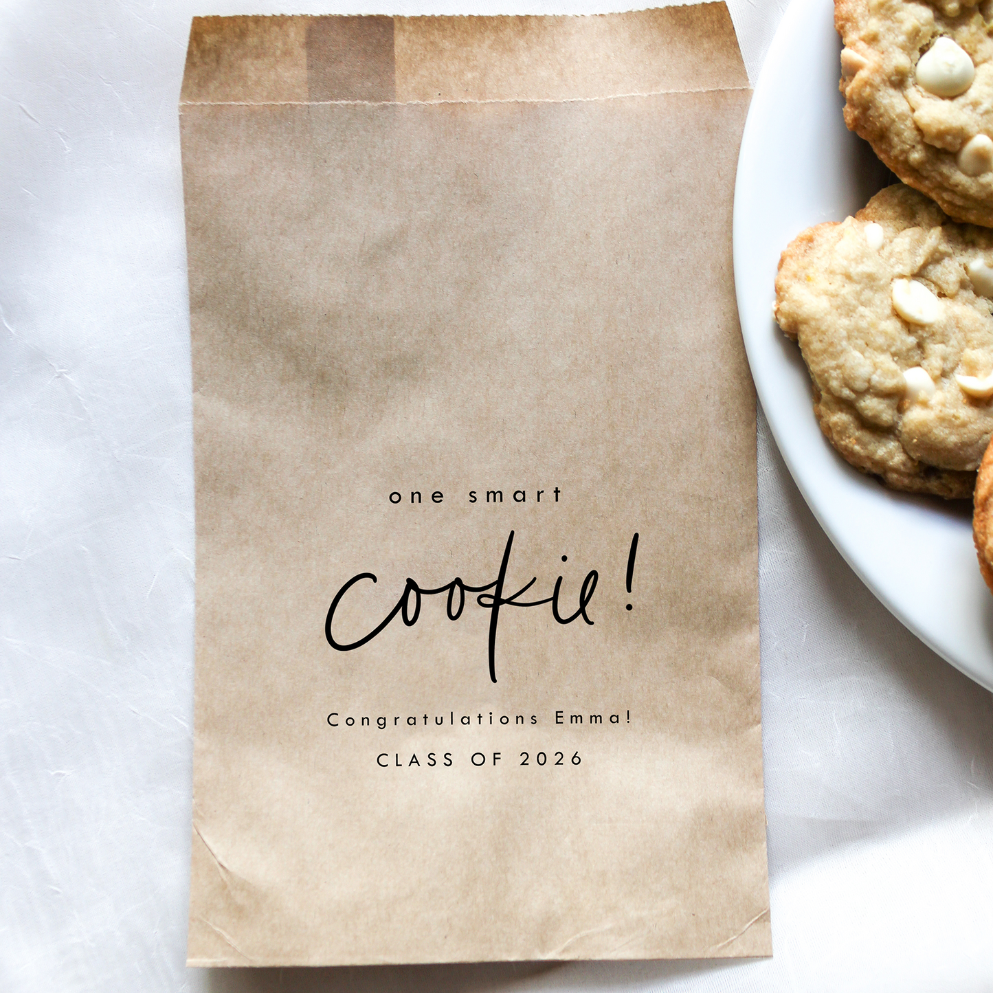 Brown paper bag with 'one smart cookie! Congratulations Emmal Class of 2026' text next to a plate of cookies on a white surface.