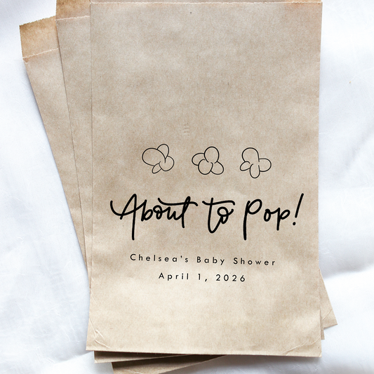 Kraft paper bags with 'About to Pop!' text and baby shower details on a white background