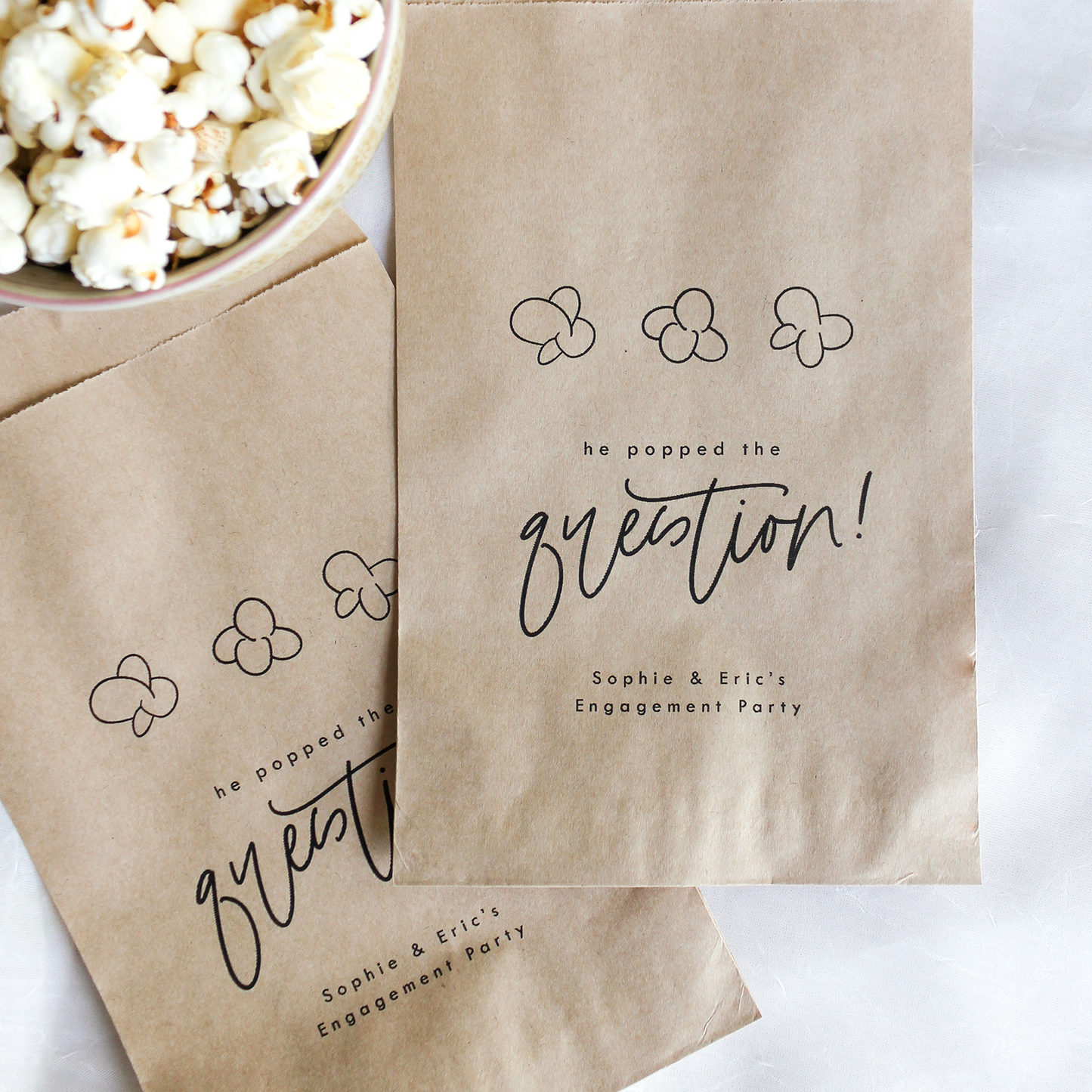 Kraft paper bags with 'he popped the question!' text next to a bowl of popcorn on a white background
