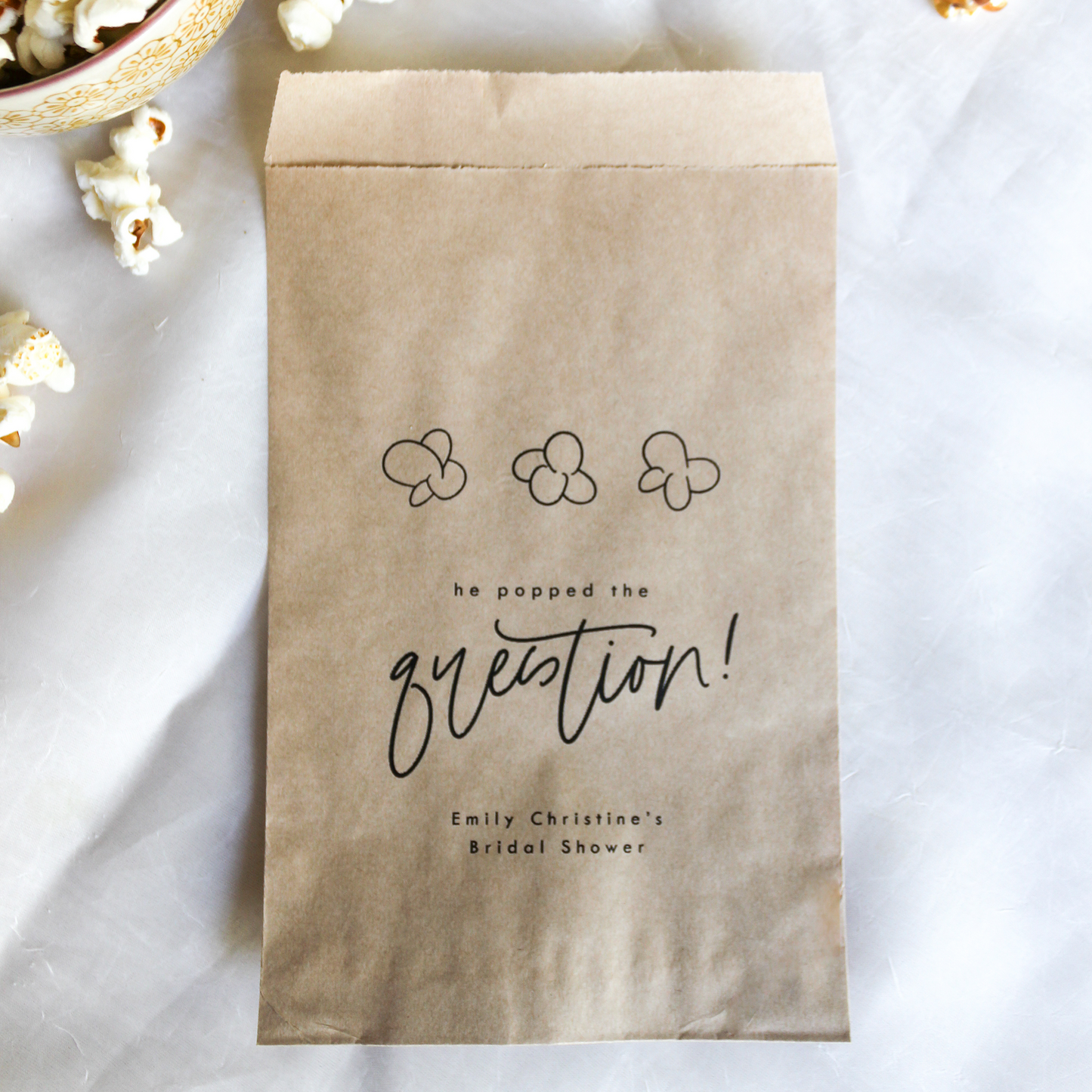 Kraft paper bag with 'he popped the question!' text on a white surface with popcorn.