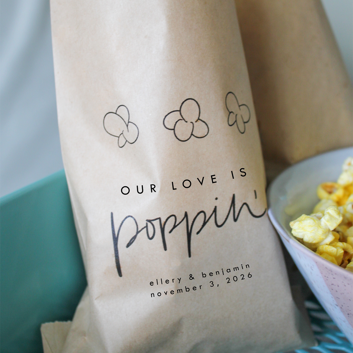 Kraft paper popcorn bag with 'Our Love is Poppin'' text and personalized details.