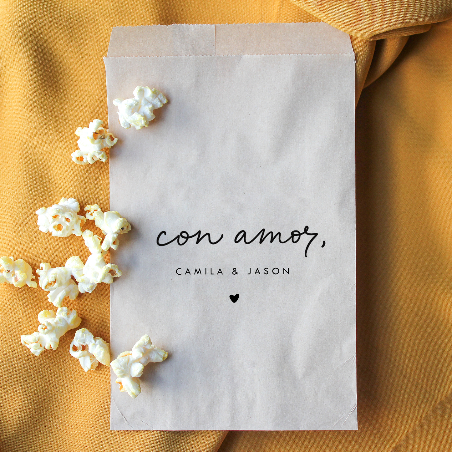 Paper bag with 'con amor, Camila & Jason' text and popcorn on a mustard yellow background