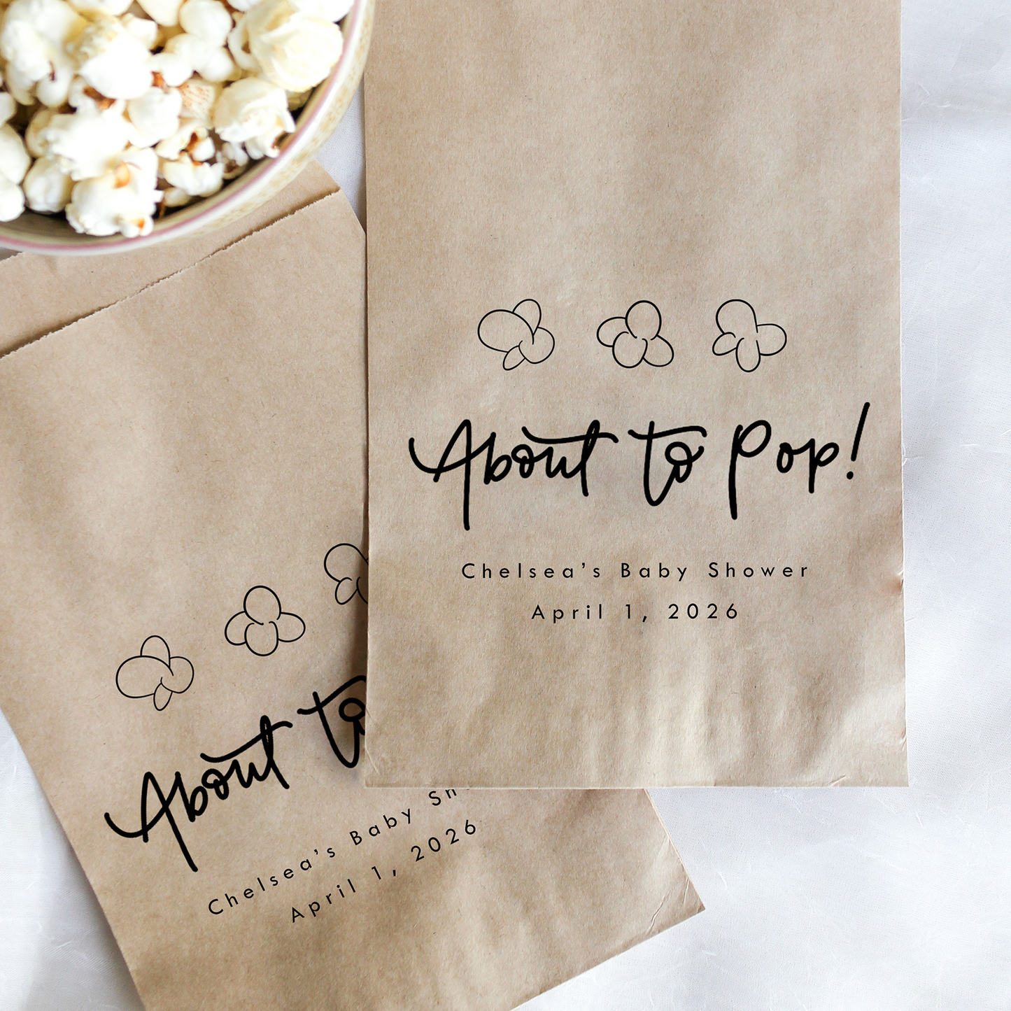 Kraft paper popcorn bags with 'About to Pop!' text and baby shower details, next to a bowl of popcorn.