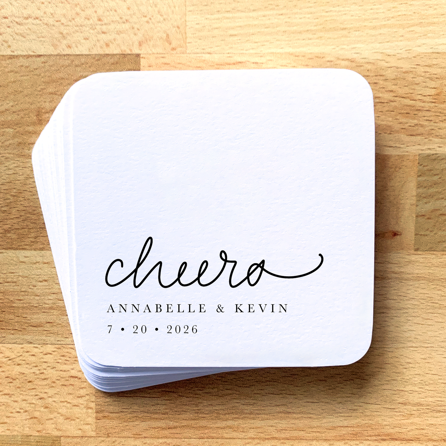 Stack of white coasters with 'cheers' and personalized text on a wooden surface