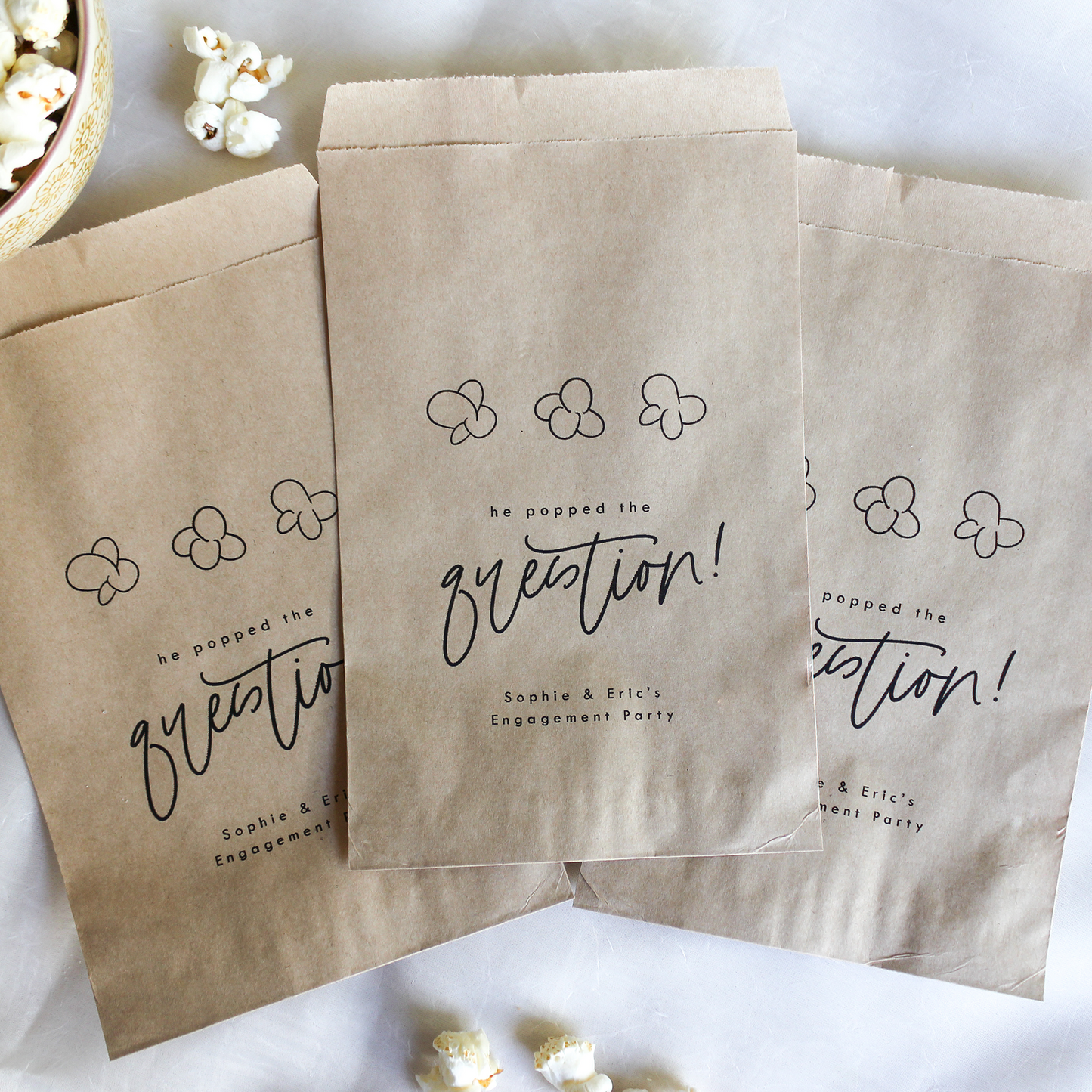 Three brown paper bags with personalized text on a white surface with popcorn.