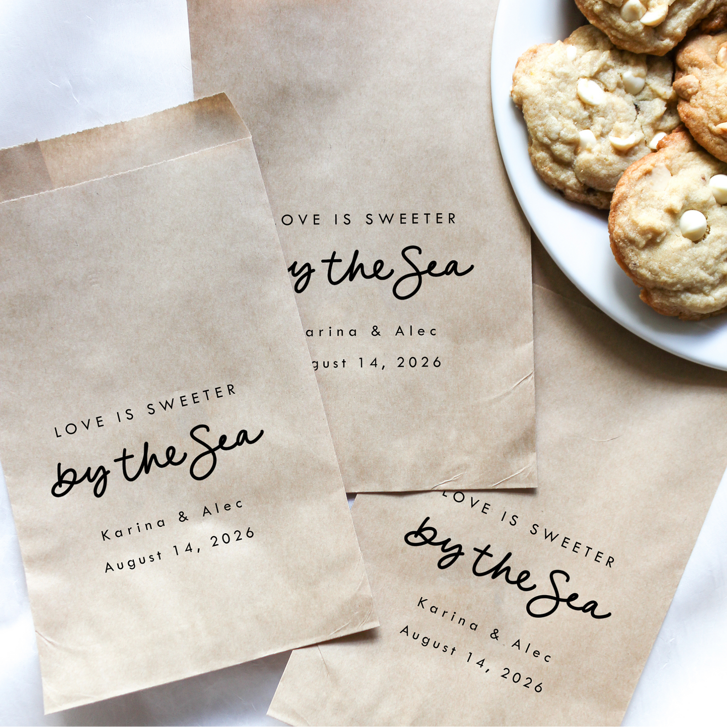 Personalized brown paper bags with 'Love is Sweeter by the Sea' text, next to a plate of cookies.