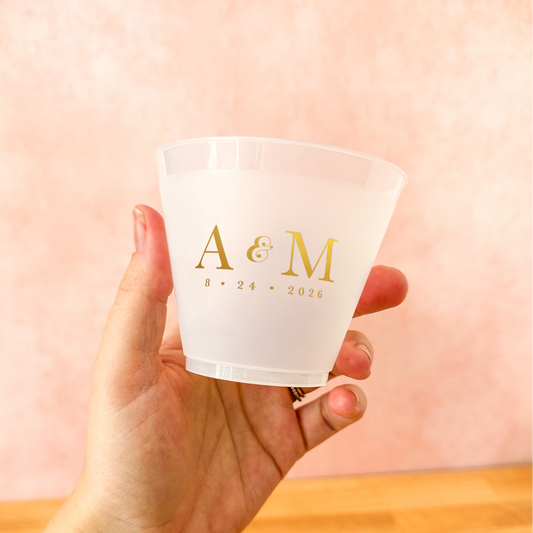 Hand holding a clear plastic cup with gold lettering against a pink background