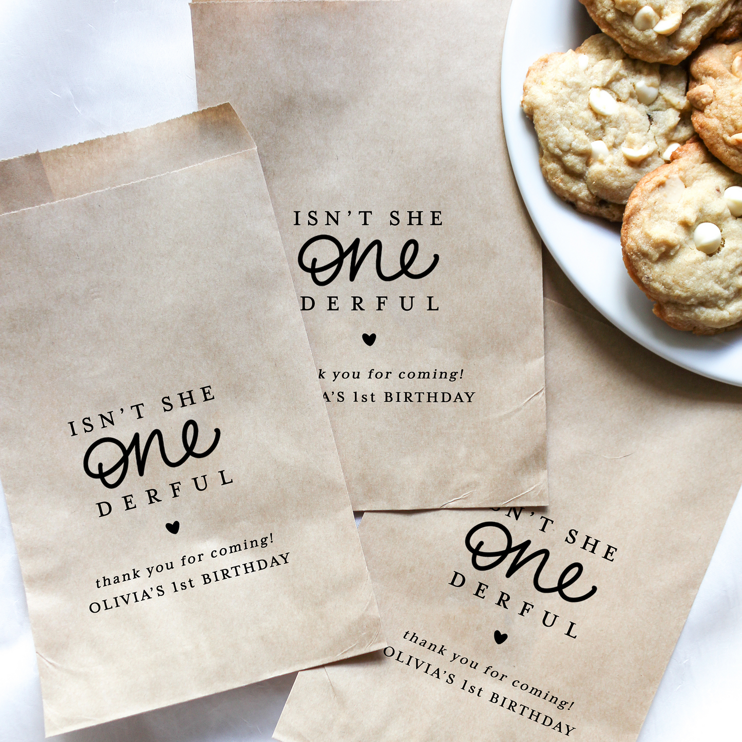 Brown paper bags with personalized text for a birthday party, next to a plate of cookies.