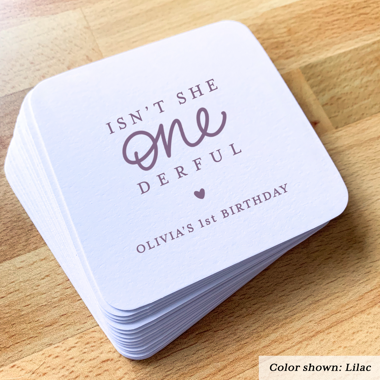 Stack of birthday coasters with text 'Isn't she one derful Olivia's 1st Birthday' on a wooden surface.
