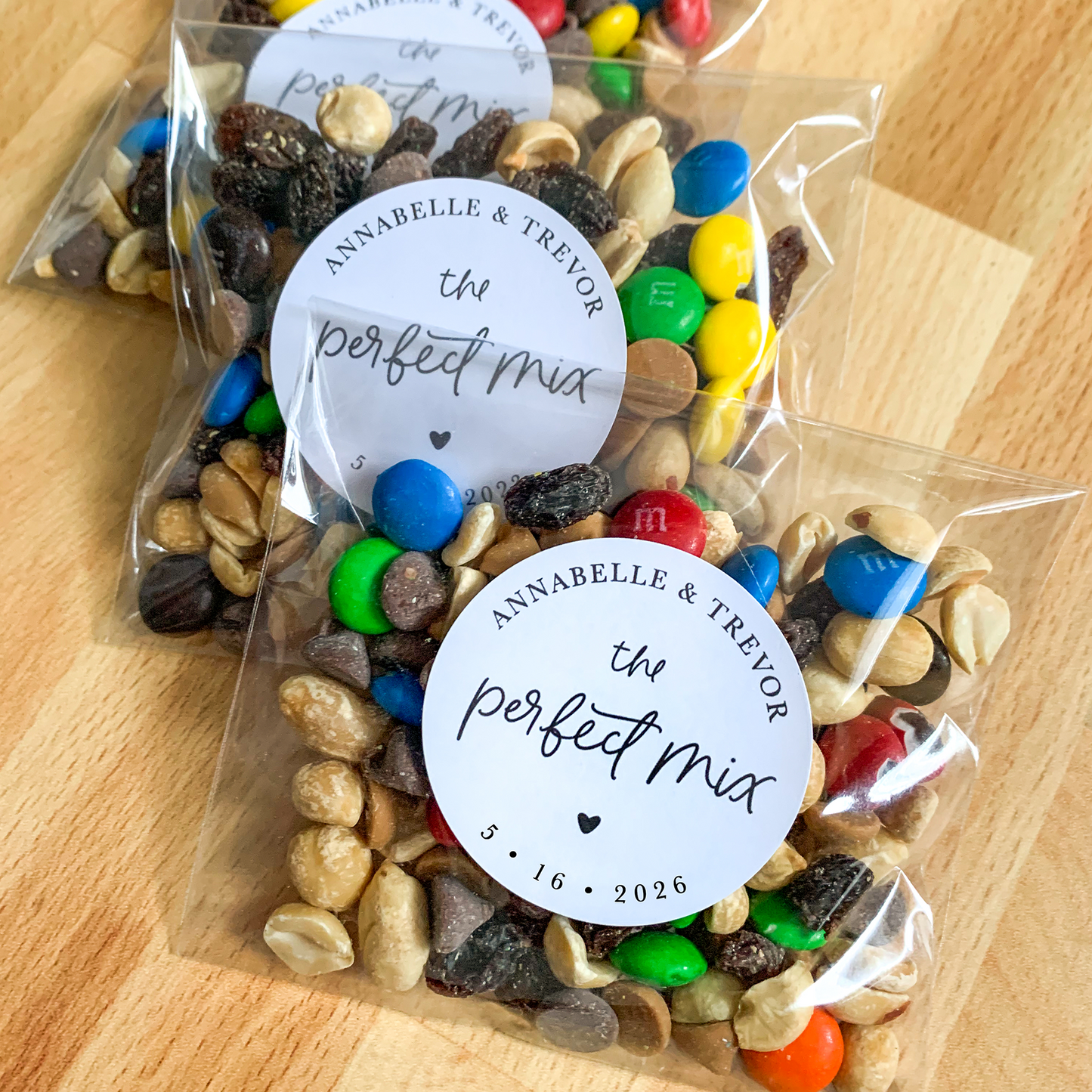 Two clear bags of trail mix with colorful candies and personalized labels on a wooden surface.